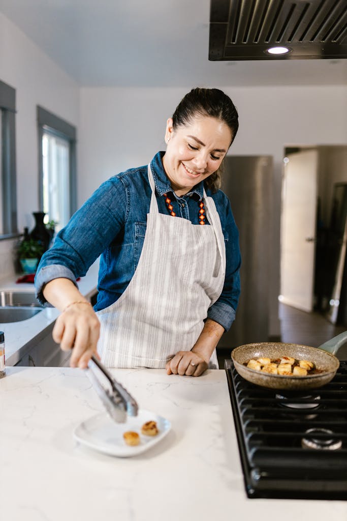 A cheerful female chef transferring food from a pan to a plate in a modern kitchen setting.