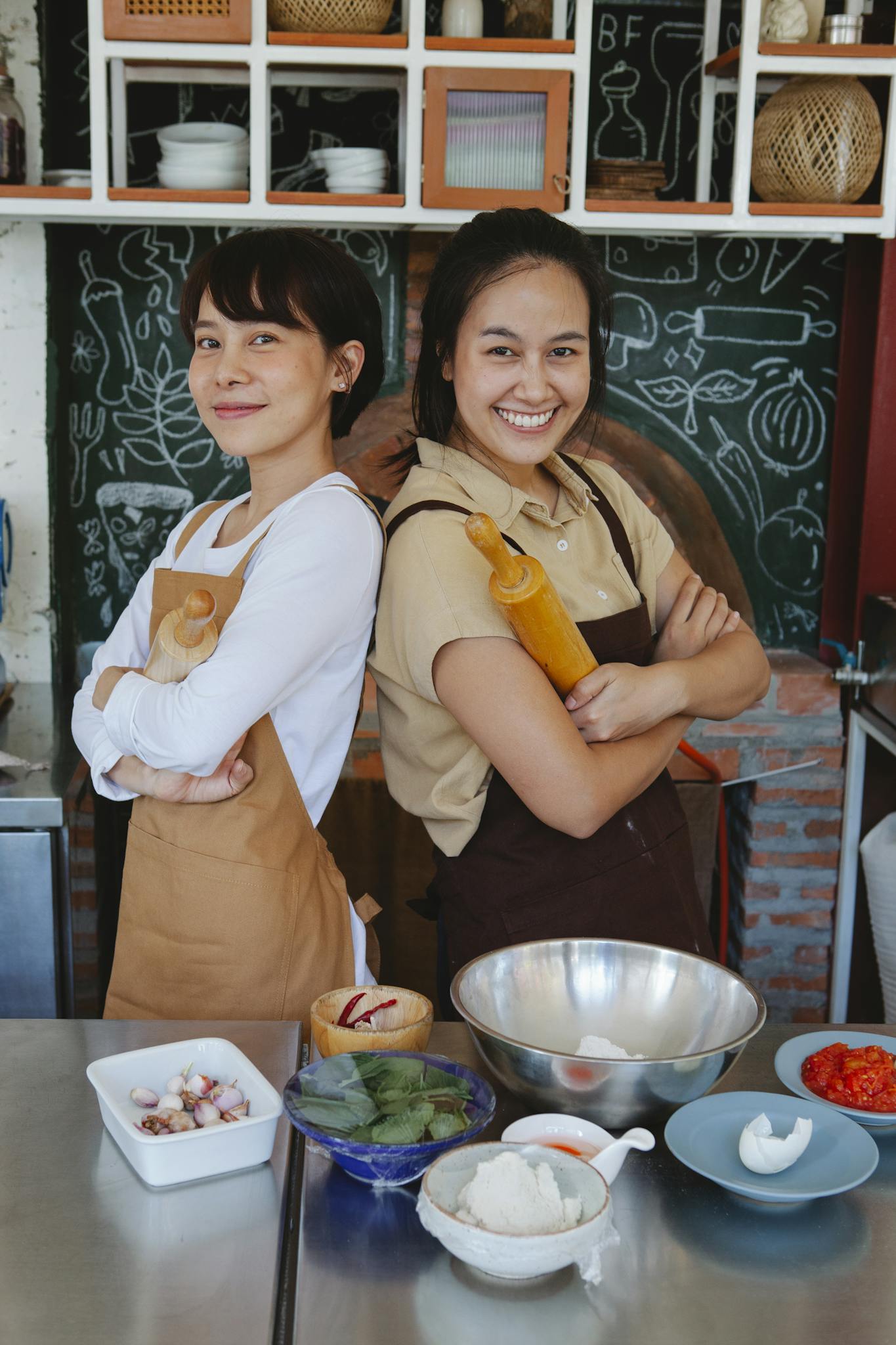 Two women chefs smile confidently in a kitchen, holding rolling pins and surrounded by ingredients.